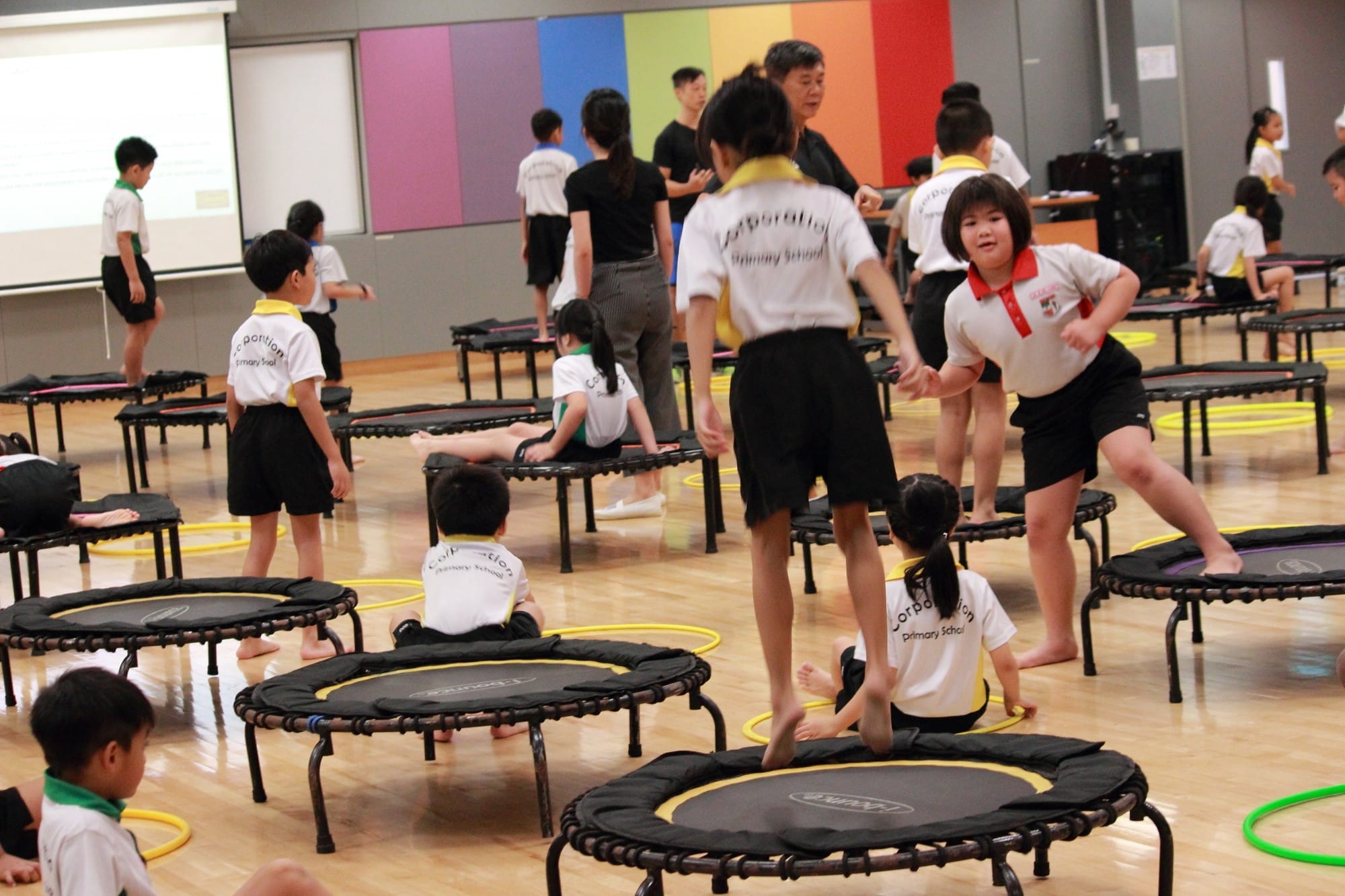 Primary school children in uniforms use small trampolines and hula hoops in a colorful indoor area.